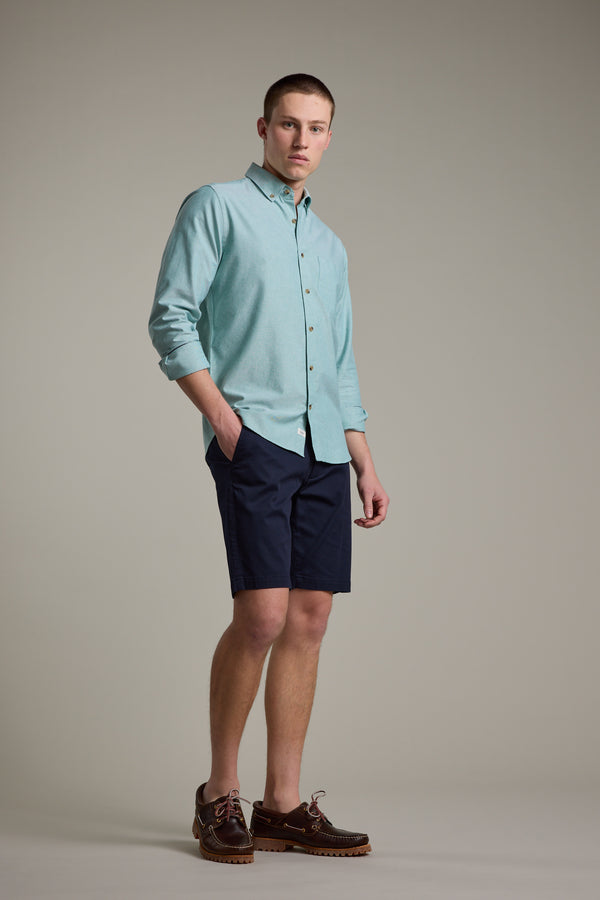 A young man stands against a plain background, confidently looking at the camera with his hands in his pockets. He wears the Barkers Thatcher Oxford Shirt in light blue, paired with navy shorts and brown boat shoes.