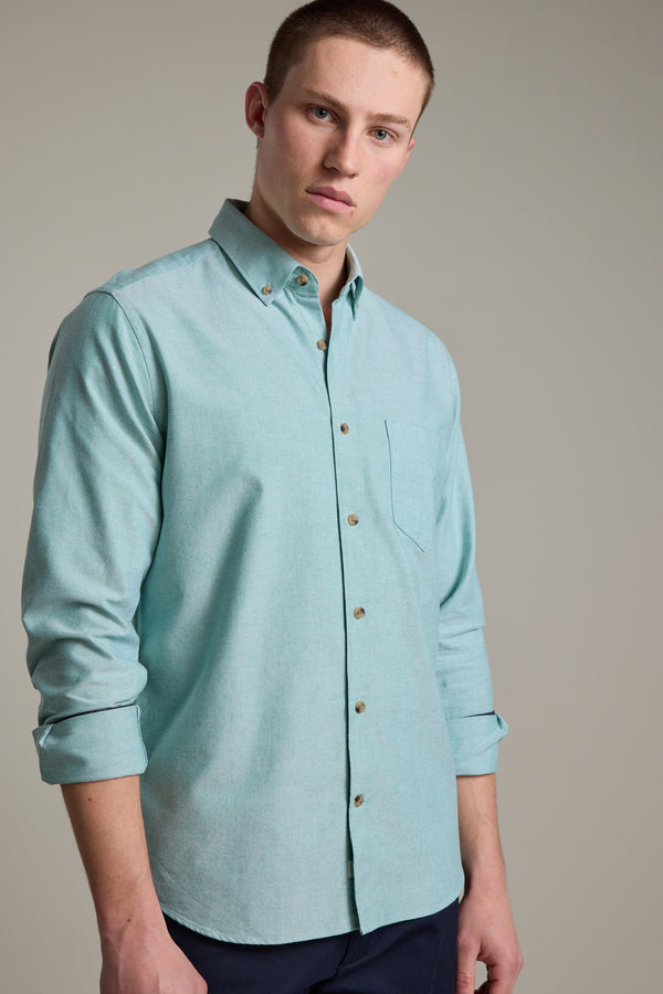 A young man in a tailored-fit, light teal Barkers Thatcher Oxford Shirt with rolled-up sleeves stands against a plain background, looking at the camera with a neutral expression. The shirt is made from responsibly sourced cotton.