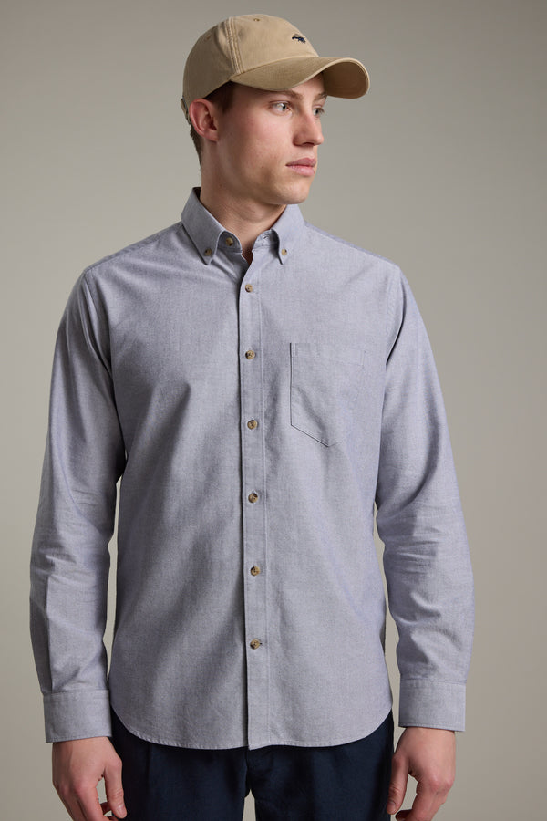 A man wearing the Barkers Thatcher Oxford Shirt in light gray and a beige baseball cap stands against a plain background, looking to his left with a neutral expression. The shirt is made from responsibly sourced cotton.