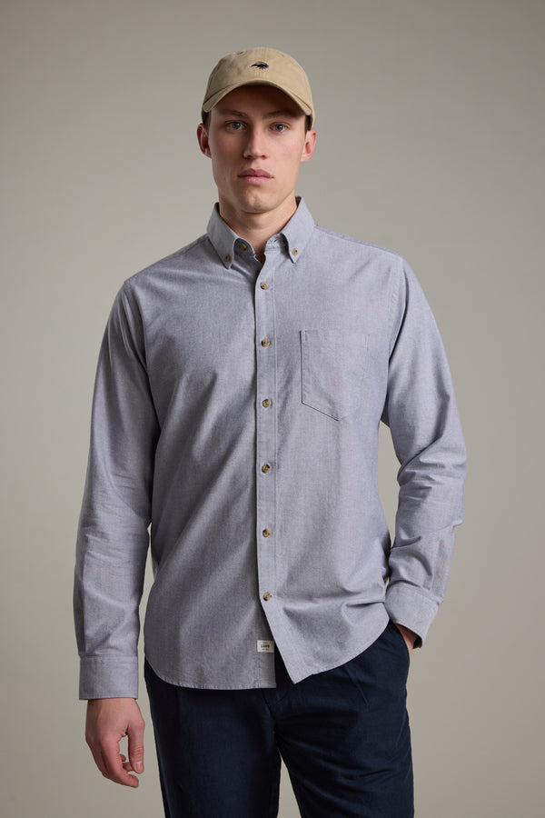 A man in a Barkers Thatcher Oxford Shirt—light gray, tailored fit, chest pocket, responsibly sourced cotton—and a tan baseball cap stands against a plain light background, facing forward with a neutral expression.