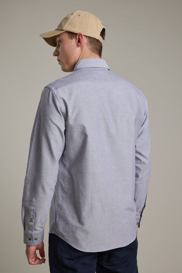 A man in a beige cap, dark pants, and a Barkers Thatcher Oxford Shirt—light gray, tailored fit, made from responsibly sourced cotton—stands facing away and looks over his shoulder against a plain background.