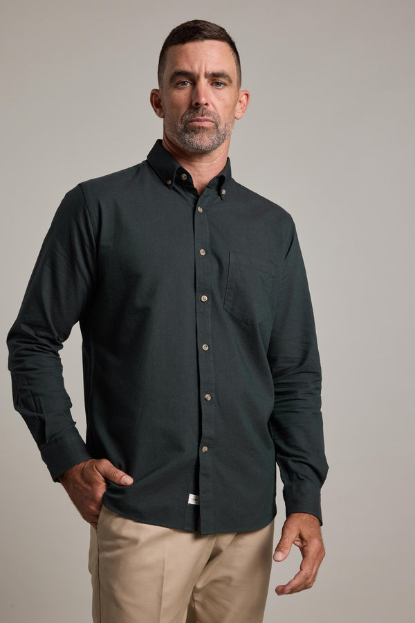 A man with short dark hair and a trimmed beard wears the Barkers Thatcher Oxford Shirt in dark green, made from responsibly sourced cotton with a chest pocket, paired with beige pants, against a plain light background.