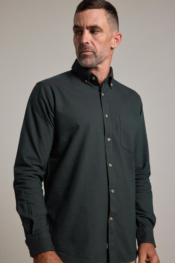 A man with short dark hair and a trimmed beard wears the Barkers Thatcher Oxford Shirt, a dark green tailored-fit with front pocket, crafted from responsibly sourced cotton. He looks left against a plain, light background.