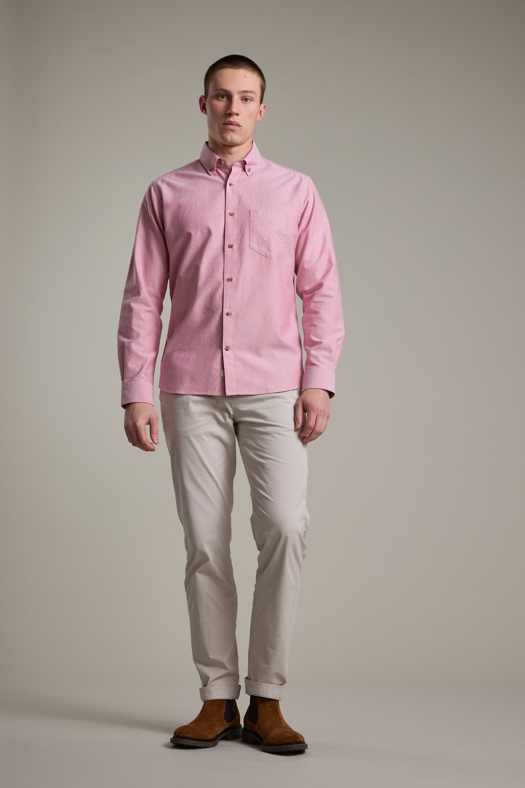 A man with short hair and a neutral expression stands against a plain background wearing the Barkers Thatcher Oxford Shirt in light pink (100% cotton), paired with light beige tailored fit pants and brown boots.