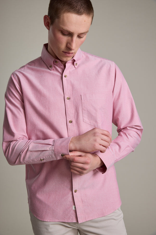 A young man with short hair adjusts his sleeve while wearing the Barkers Thatcher Oxford Shirt, a light pink tailored-fit shirt made from responsibly sourced cotton, against a plain background.