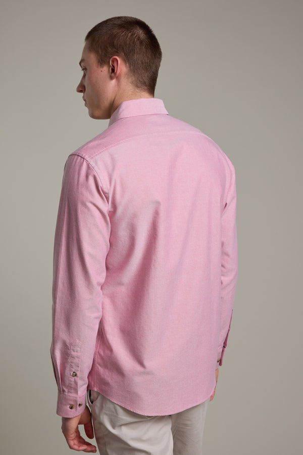 A man stands with his back to the camera, wearing Barkers' Thatcher Oxford Shirt in light pink, crafted from responsibly sourced cotton, paired with light-colored pants against a plain, neutral background.