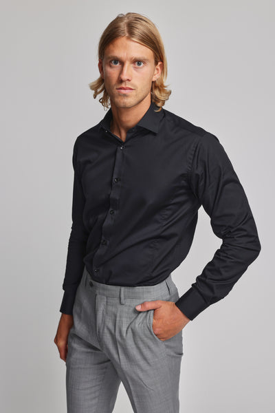 A man with shoulder-length blond hair models the Barkers Tribeca Reg Cuff Shirt, a tailored black dress shirt, paired with light gray trousers. He stands against a plain background, looking seriously at the camera with one hand in his pocket.