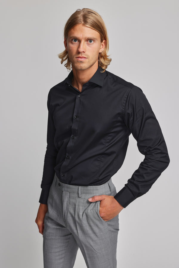A man with shoulder-length blond hair models the Barkers Tribeca Reg Cuff Shirt, a tailored black dress shirt, paired with light gray trousers. He stands against a plain background, looking seriously at the camera with one hand in his pocket.
