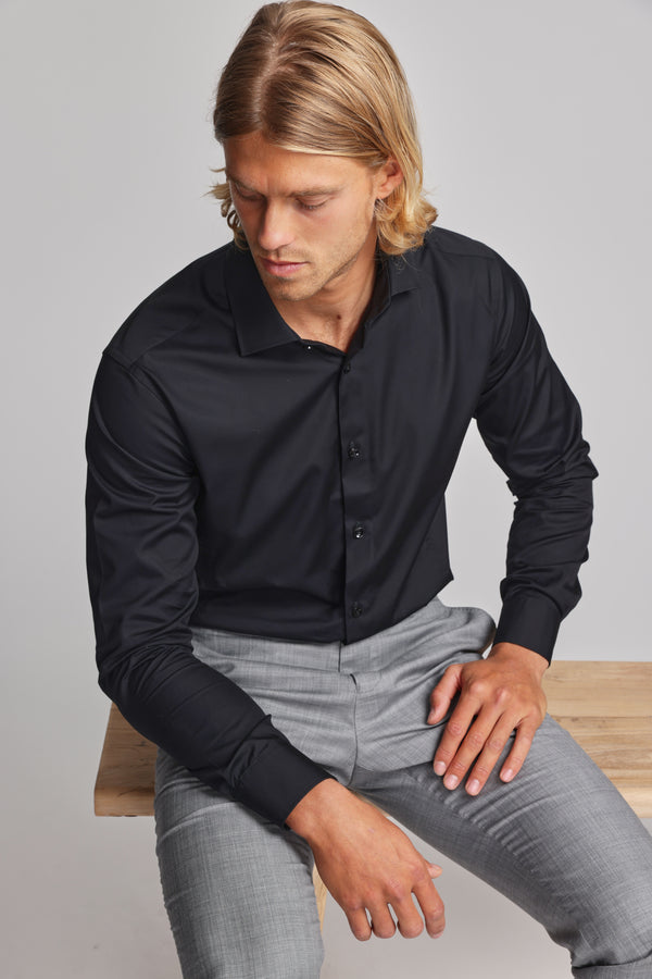 A man with shoulder-length blonde hair, dressed in a wrinkle-free black Tribeca Reg Cuff Shirt from Barkers and light gray pants, sits on a wooden bench looking down with a neutral expression.