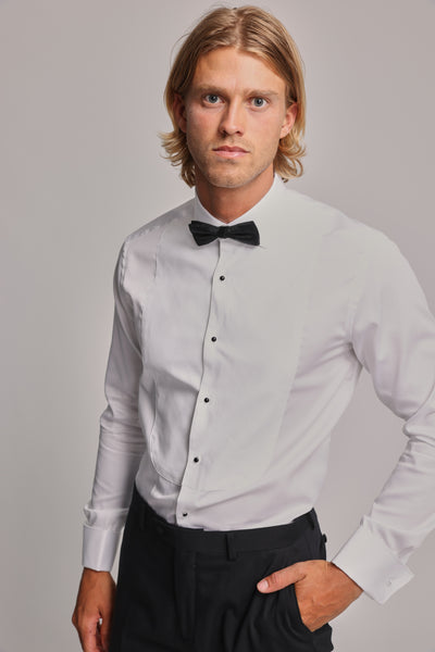 A young man with shoulder-length blonde hair models the Barkers Eldridge Dress Shirt, paired with a black bow tie and black pants. He stands confidently with one hand in his pocket, facing the camera against a plain background.