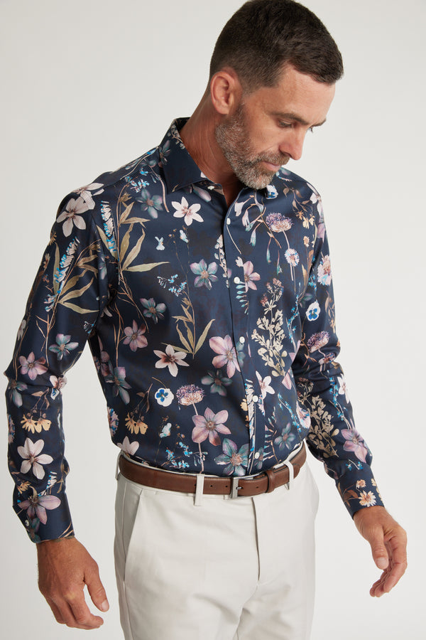 A man wearing a dark blue Barkers Apothecary AOP Business Shirt with a colorful floral pattern, paired with light pants and a brown belt. This classic fit shirt is crafted from responsibly sourced cotton.