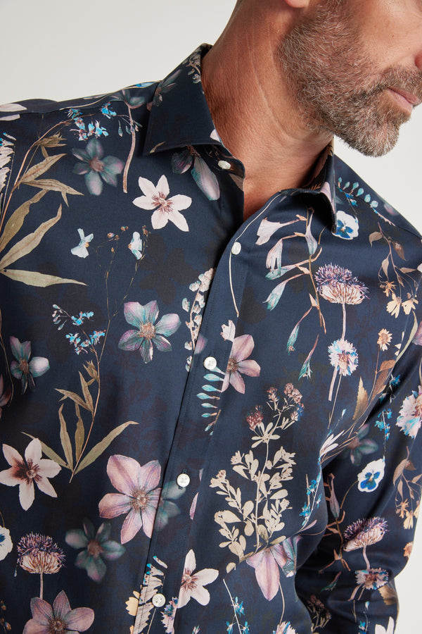 A man with a short beard wears the Barkers Apothecary AOP Business Shirt, a classic-fit, responsibly sourced cotton shirt in dark blue with a vivid floral print in pink, purple, beige, and green. Cropped at the shoulders.
