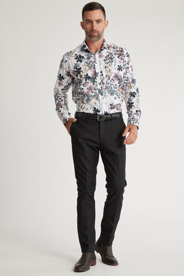 A man with short hair and a trimmed beard stands against a plain background wearing the Barkers Apothecary AOP Business Shirt, responsibly made from cotton, paired with black pants, a black belt, and dark brown dress shoes. One hand is in his pocket.