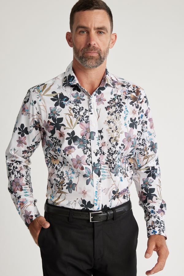 A man with short brown hair and a beard wears the Barkers Apothecary AOP Business Shirt in a colorful floral pattern with black trousers, standing against a plain light background.