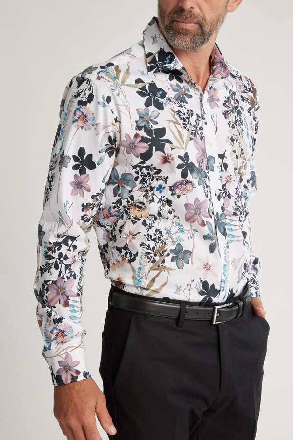 A bearded man wears a classic fit Barkers Apothecary AOP Business Shirt, crafted from responsibly sourced cotton with a colorful floral print, tucked into black pants and secured with a black belt against a plain light background.