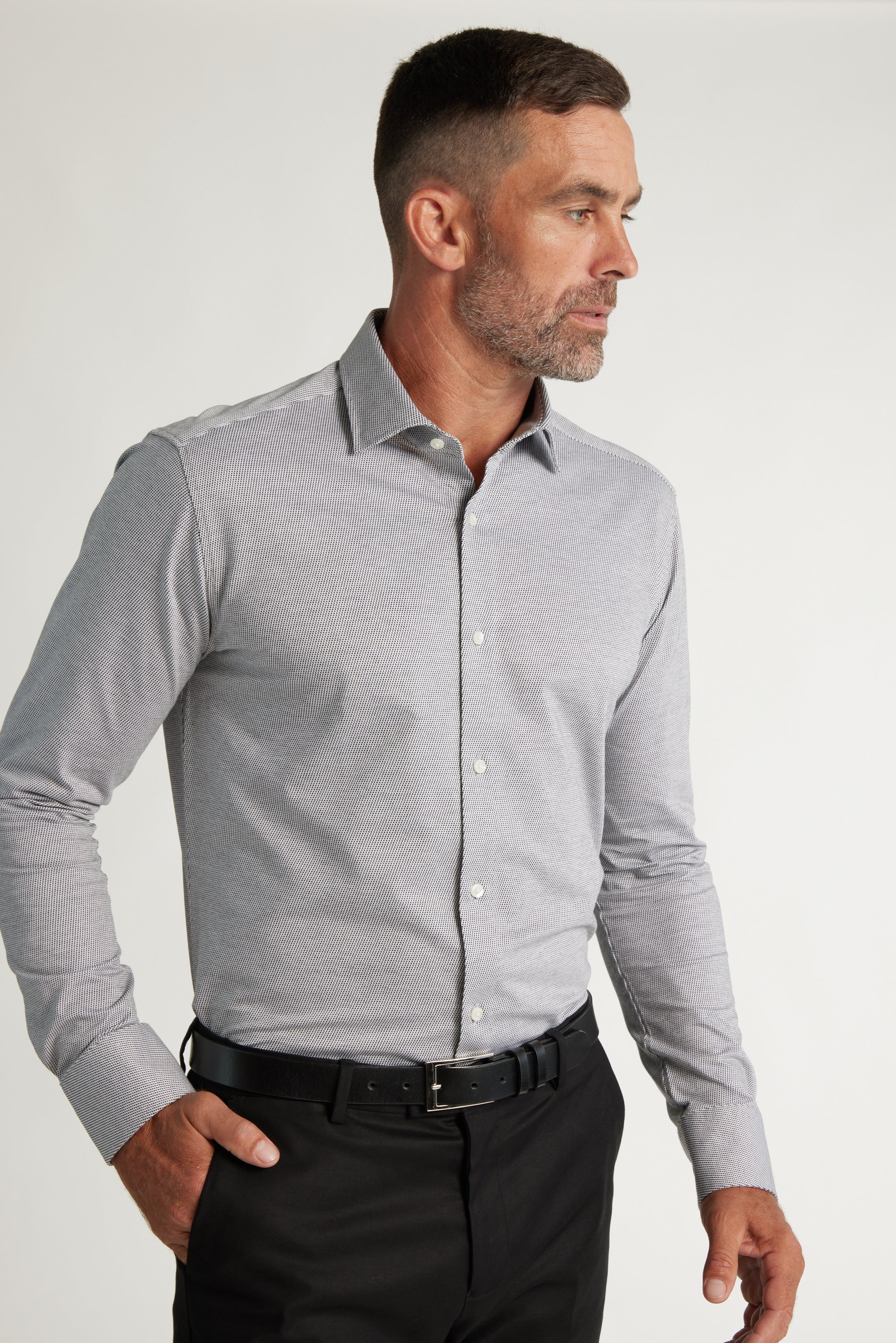 A man with short dark hair and a beard wears a Barkers Goodrich Knit Business Shirt in light gray and black trousers with a belt, standing against a plain light background and looking to his left.