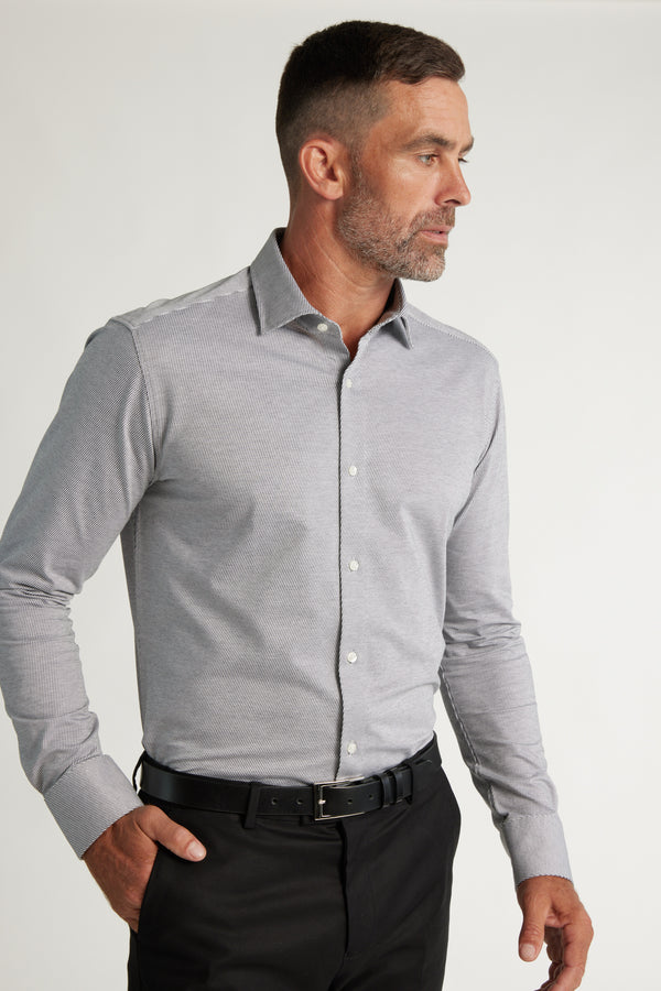 A man with short dark hair and a beard wears a Barkers Goodrich Knit Business Shirt in light gray and black trousers with a belt, standing against a plain light background and looking to his left.