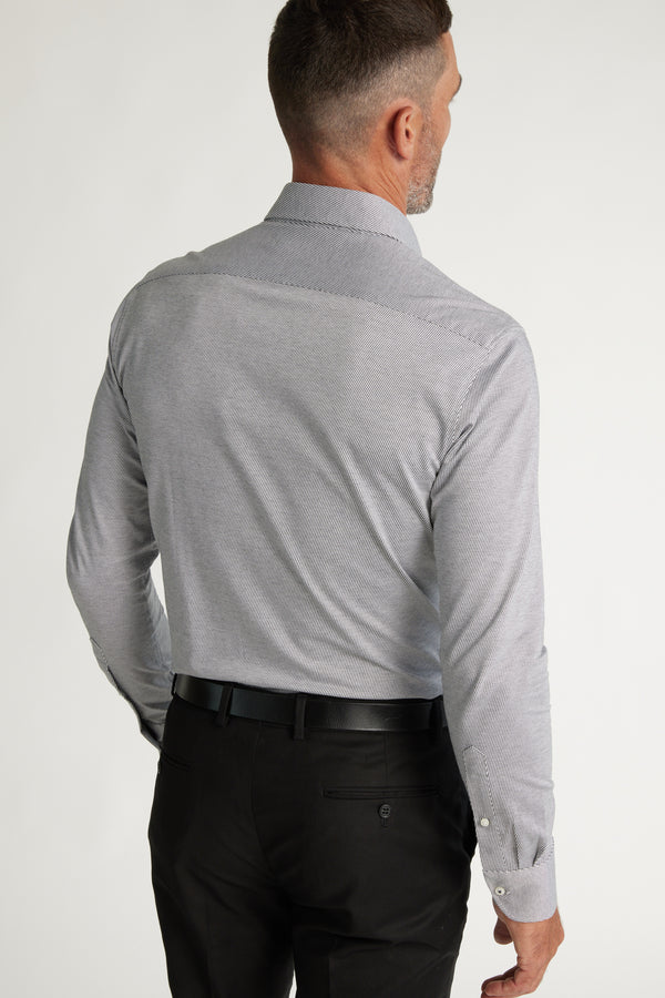 A man faces away, wearing the Barkers Goodrich Knit Business Shirt in light gray, made from responsibly sourced cotton, paired with black trousers and a black belt against a plain light background.