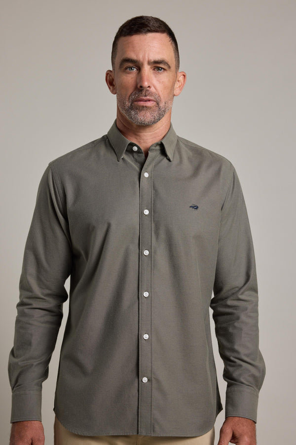 A man with short brown hair and a trimmed beard wears the Barkers Maynard Oxford Shirt in olive green, paired with tan pants. He stands against a plain light gray background, looking directly at the camera.