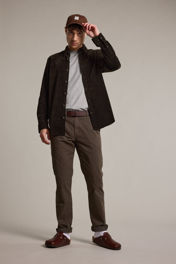 A man stands against a plain background, wearing the Barkers Washed Cotton Cap by Barkers, a dark brown button-up shirt over a gray t-shirt, brown belted pants, white socks, and brown shoes. He holds the cap’s brim with one hand.