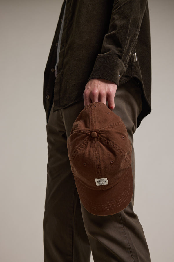 A person in a dark brown shirt and pants holds the Barkers Washed Cotton Cap by the brim, arm relaxed at their side. The minimalist background keeps the focus on the Barkers cap.