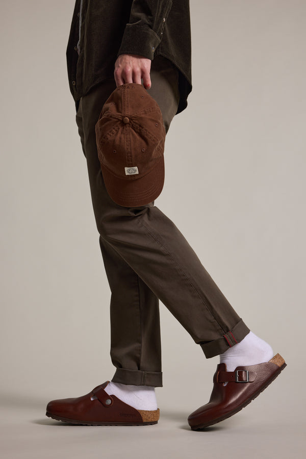 A person in brown pants, white socks, and brown buckle shoes holds a Barkers Washed Cotton Cap by Barkers, standing against a plain background. The photo is cropped to show only the lower body.