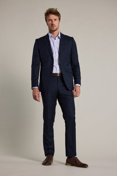 A man faces forward against a light background, wearing Barkers Wiltshire Suit Trousers in dark navy merino wool, paired with a white dress shirt, brown belt, and brown dress shoes.