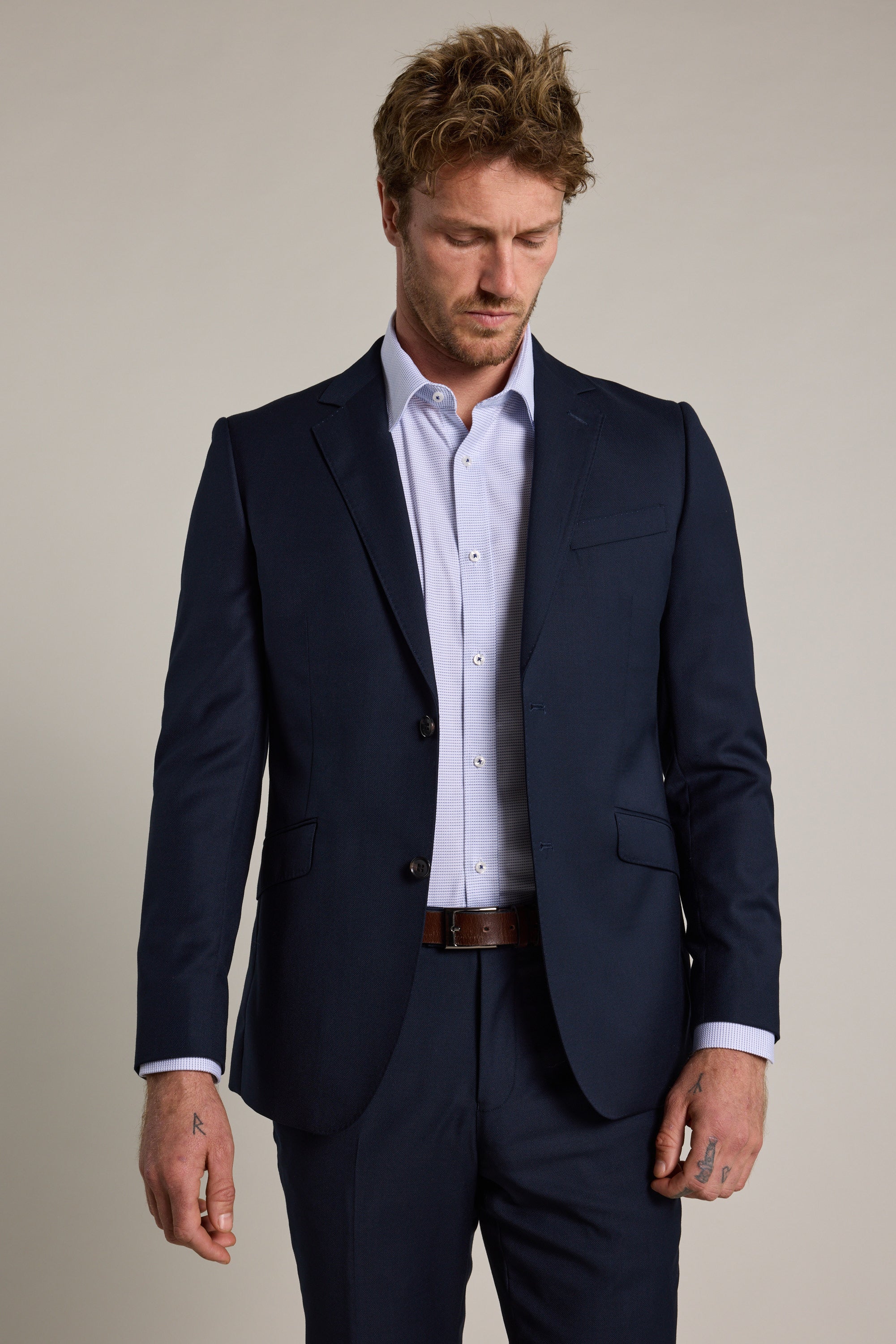 A man with tousled blond hair models the Barkers Wiltshire Suit Jacket, a navy single-breasted style, over a light blue dress shirt against a plain background. His hands rest by his sides as he looks down with a neutral expression.