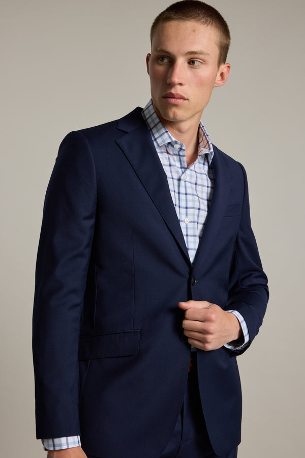 A young man in a Barkers Wesley Check Business Shirt stands against a plain background, wearing a navy blue suit and looking slightly left with a neutral expression.