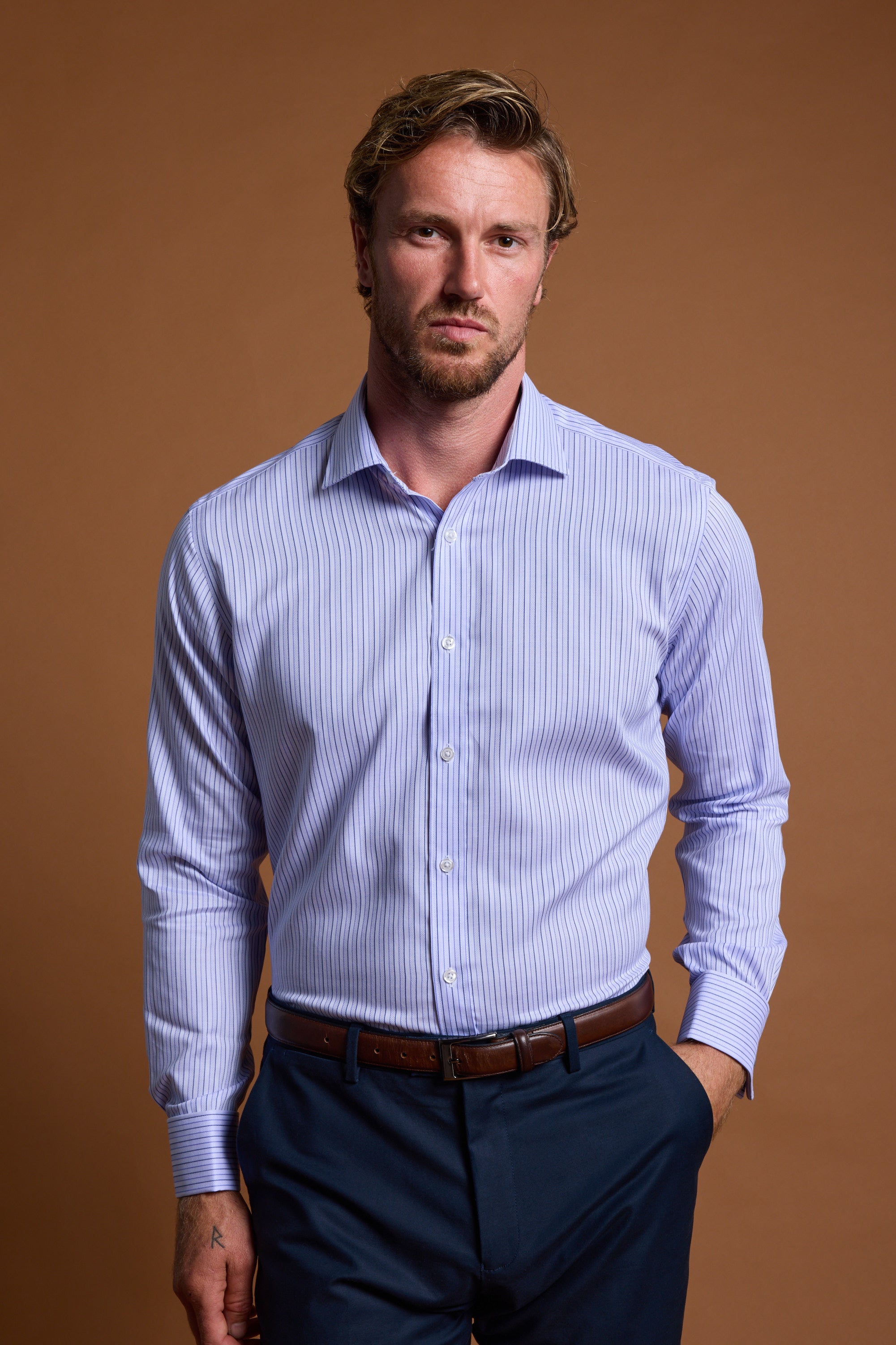 A man with light brown hair and a beard wears the Barkers Asmara French Cuff Business Shirt paired with dark trousers, posing against a plain brown background with one hand in his pocket and looking at the camera.