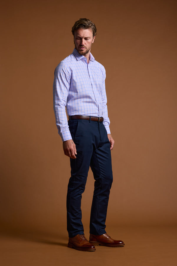 A man with short, wavy hair stands against a plain brown background wearing the Barkers Larry Check Business Shirt—light blue and wrinkle-free—paired with dark navy trousers, a brown belt, and matching dress shoes. He looks slightly to his left.