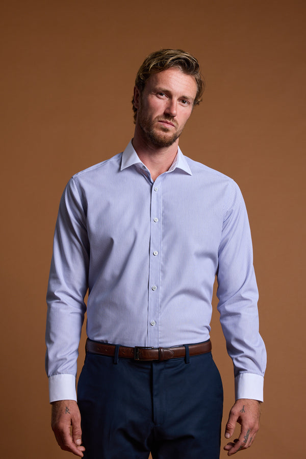 A man with wavy light brown hair and a beard stands against a plain brown background, wearing the Barkers Felix Stripe Business Shirt (tailored fit, Eco Easy Care fabric), dark blue trousers, and a brown belt with a neutral expression.
