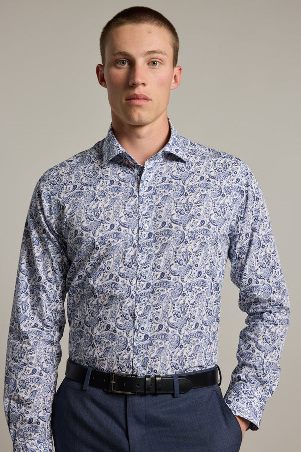 A young man with short hair wears the Buckman Print Business Shirt by Barkers, featuring a blue and white paisley pattern, paired with navy trousers and a black belt, standing against a plain light gray background.