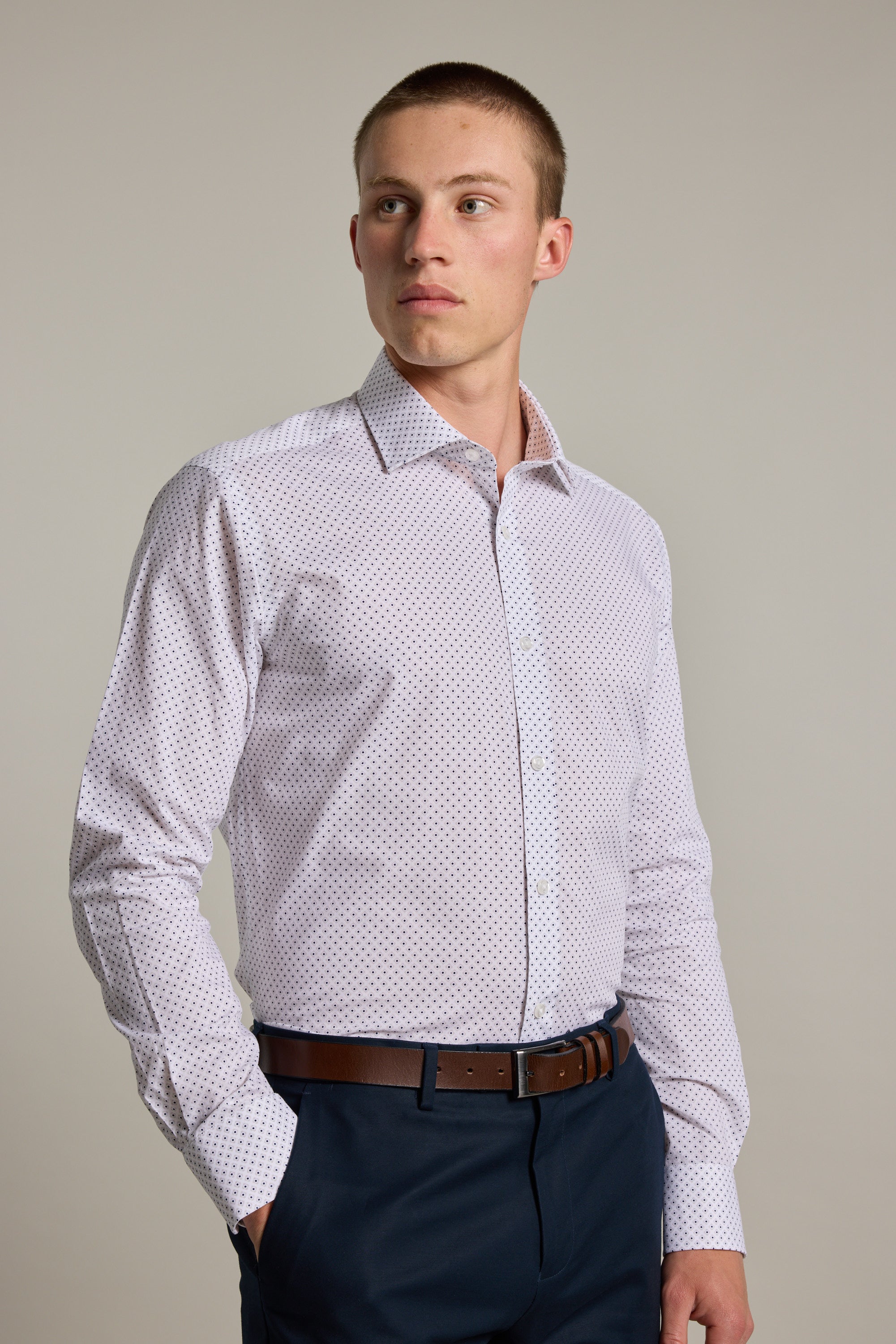 A young man with short hair wears the Barkers Foster Print Business Shirt in a tailored fit, tucked into dark pants with a brown belt. He stands against a plain background, looking slightly to his left.