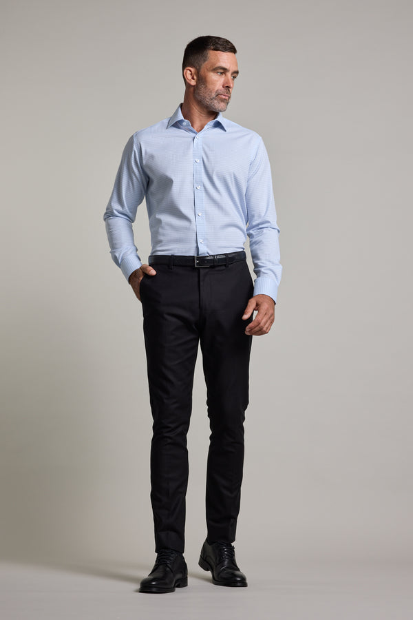 A man stands against a plain background, wearing the Barkers Hunter Check Business Shirt, dark slim-fit trousers, a black belt, and black dress shoes. He has one hand in his pocket and looks to the side.