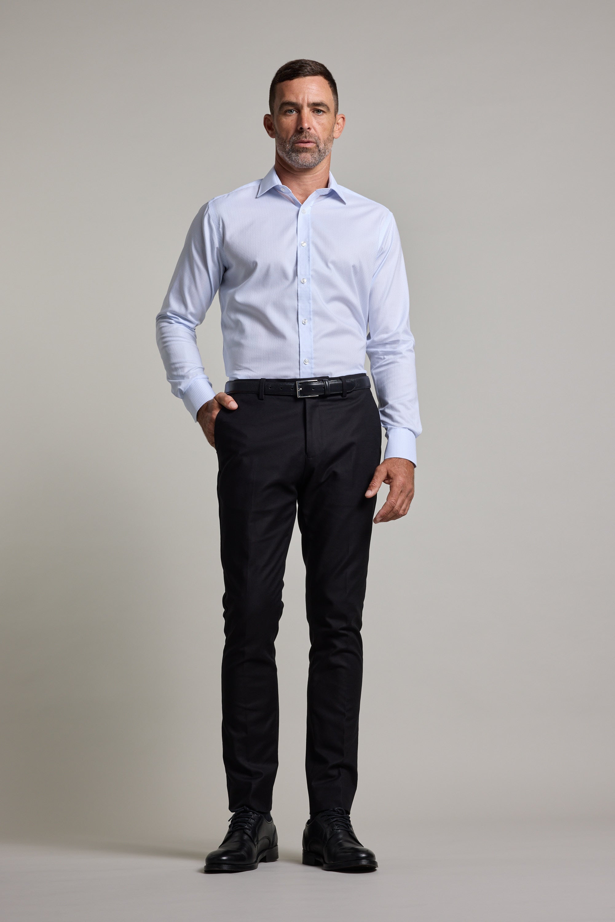 A man faces forward in a tailored fit Barkers Penny Texture Business Shirt, paired with dark dress pants and black shoes, one hand in his pocket against a plain light background.
