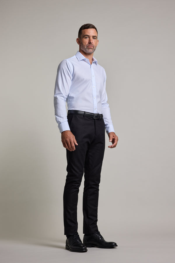 A man with short hair and a trimmed beard stands against a plain light background, wearing the Barkers Penny Texture Business Shirt in light blue, black trousers, a black belt, and dress shoes. He has a neutral expression with his hands at his sides.