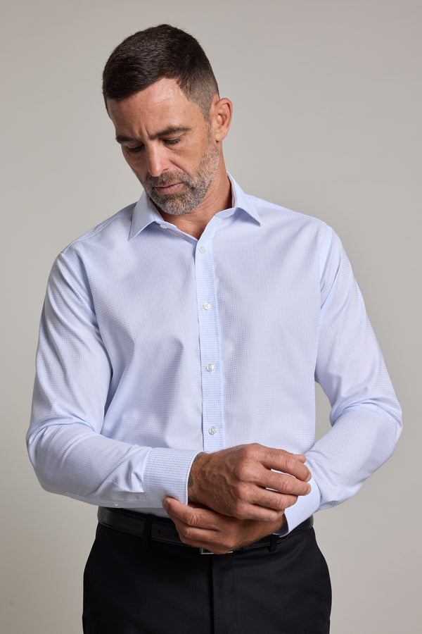 A man with short dark hair and a beard, wearing the Barkers Penny Texture Business Shirt in light blue and black pants, looks down as he adjusts his left cuff against a plain, neutral background.