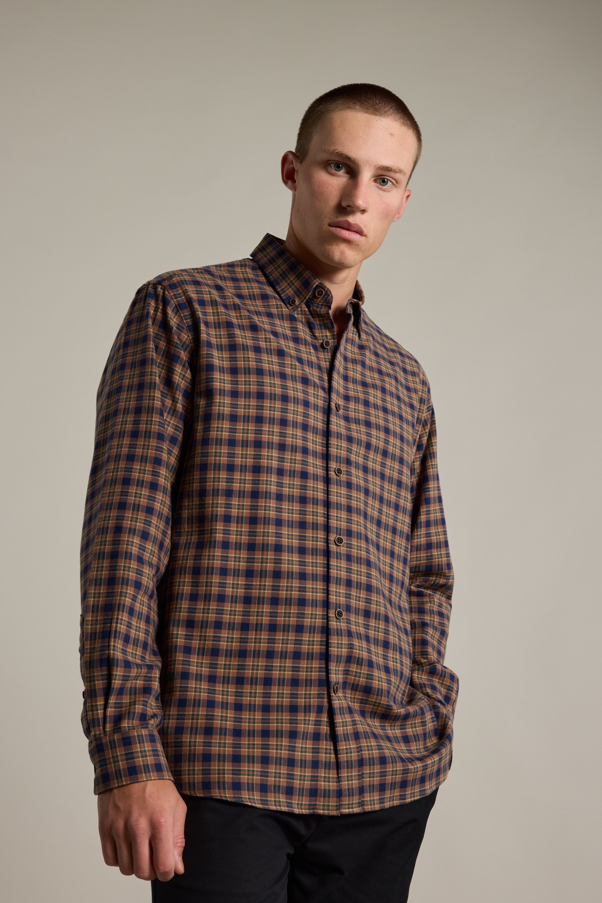 A young man with short hair wears the Barkers Manselheck Shirt in brown and navy, made from responsibly sourced cotton. He stands against a plain background with one hand in his pocket, looking at the camera.
