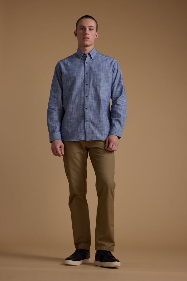 A young man stands against a tan background, wearing the Barkers Blaine Stripe Shirt in a classic fit with blue stripes, khaki pants, and dark sneakers. He faces forward with a neutral expression and relaxed posture.