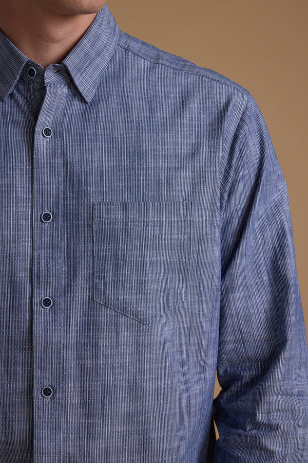 A person wears the Barkers Blaine Stripe Shirt, a blue, classic fit top crafted from responsibly sourced cotton, shown from shoulders to mid-torso against a beige background.