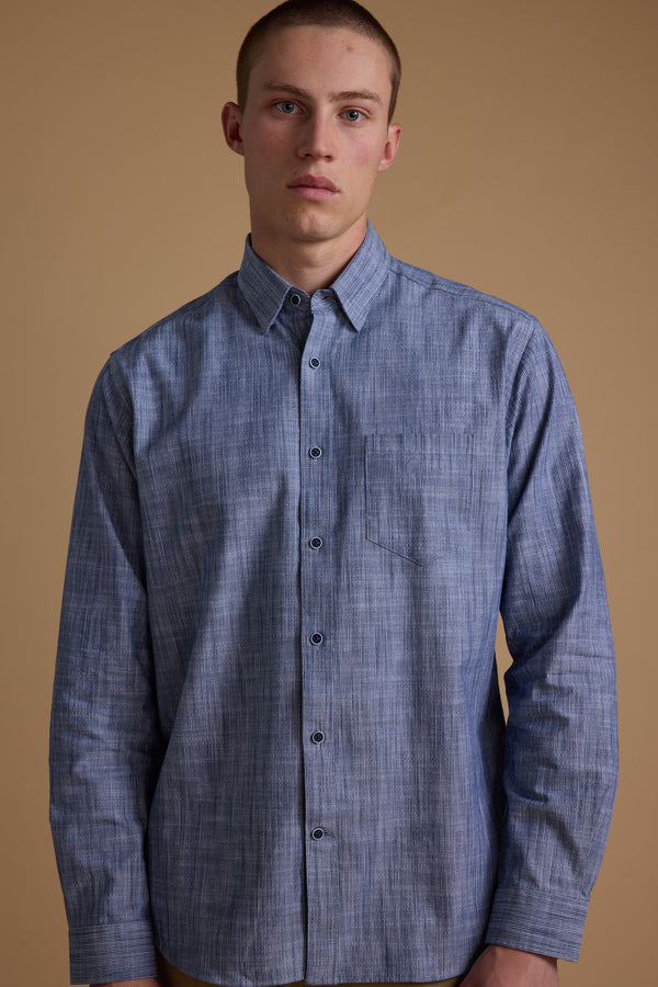 A young man with short hair wears the Barkers Blaine Stripe Shirt, a classic fit made from responsibly sourced cotton, featuring blue stripes. He stands against a plain beige background, looking directly at the camera with a neutral expression.