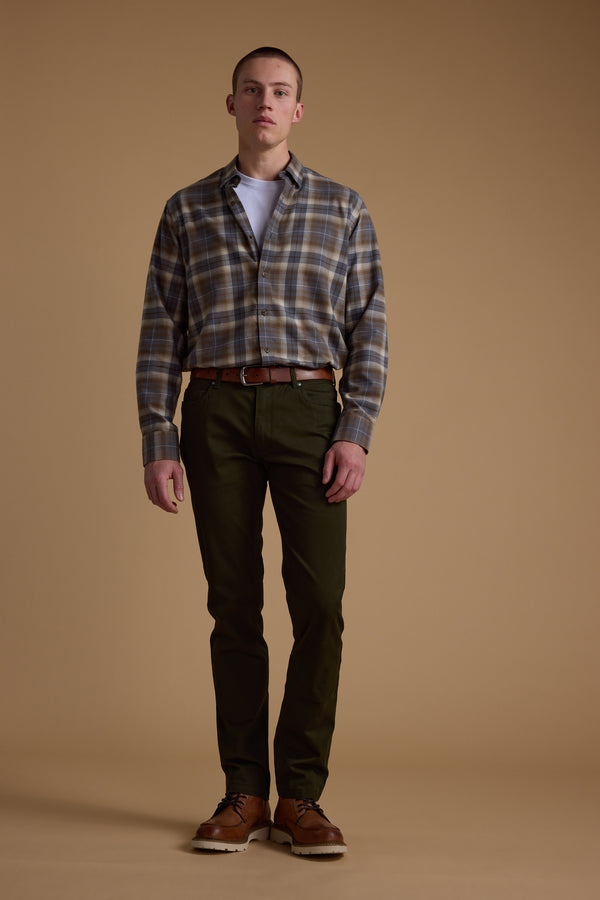 A man stands facing forward against a plain tan background, wearing a Barkers Derick Check Shirt in gray and brown plaid over a white t-shirt, with olive green pants, brown belt and shoes—made from responsibly sourced cotton.