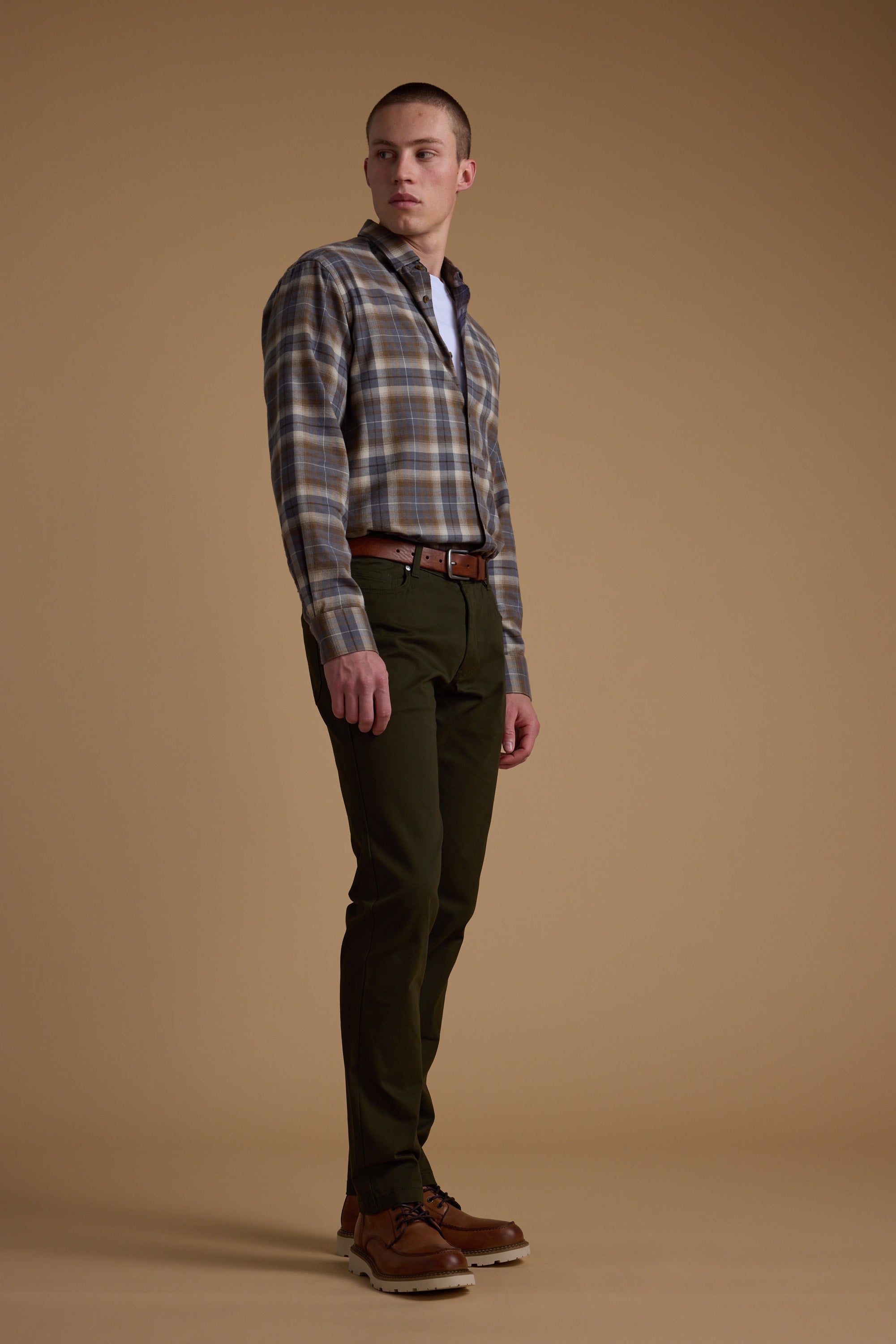 A man stands against a plain brown backdrop wearing a plaid shirt, Barkers Beckett 5-Pocket Pant, a brown belt, and brown shoes. He looks slightly to the side with a neutral expression and relaxed posture.