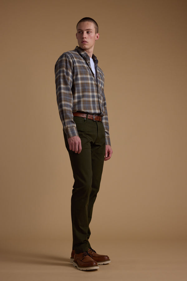 A man wearing a Barkers Derick Check Shirt stands against a plain brown background with dark green pants, a brown belt, and brown shoes. He gazes slightly to the side. The shirt is crafted from responsibly sourced cotton.