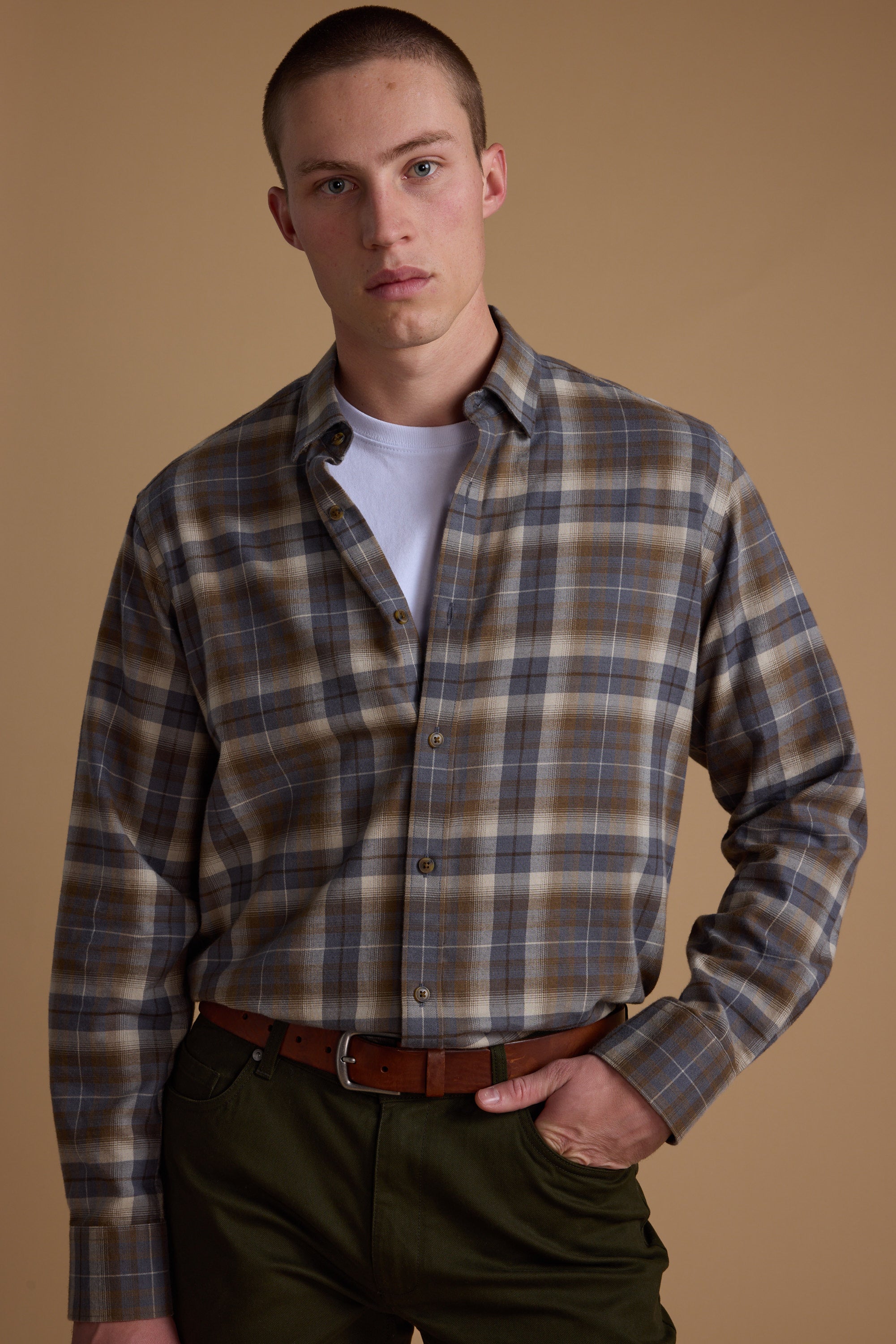 A young man with short hair and a neutral expression stands against a beige background wearing the Barkers Derick Check Shirt, made from responsibly sourced cotton, over a white tee. He pairs it with dark green pants and a brown belt.