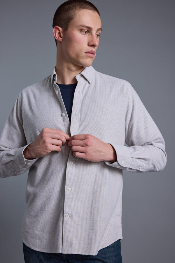 A young man with short hair wears the Barkers Maynard Oxford Stripe Shirt, made from responsibly sourced cotton, over a dark tee. He stands against a plain gray background, looking left and fastening a button on his shirt.
