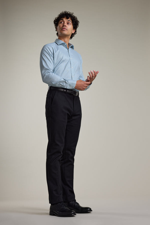 A man with curly hair models the Barkers Zione Business Shirt, pairing it with black trousers and shoes. He stands against a plain background, looking upward with one hand raised and the other by his waist.