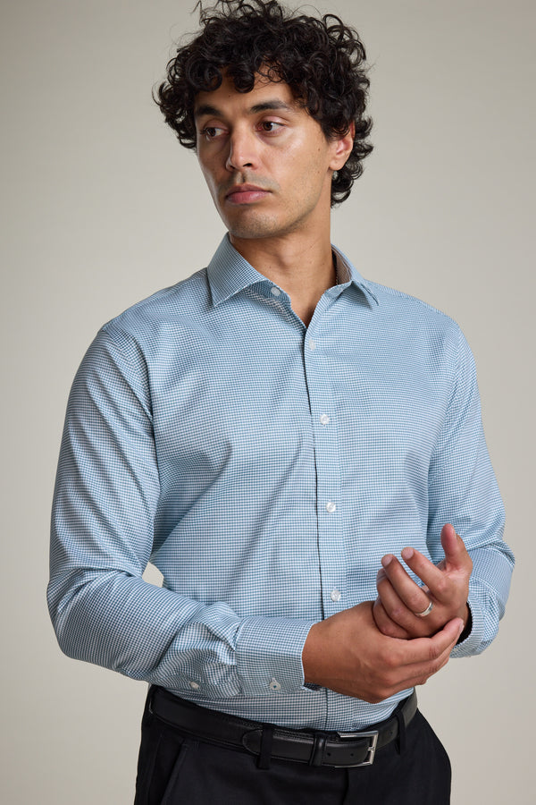 A man with curly dark hair wears the Barkers Zione Business Shirt, a light blue houndstooth-patterned shirt, and black pants. He stands against a plain background, looking left while adjusting his right sleeve with his left hand.