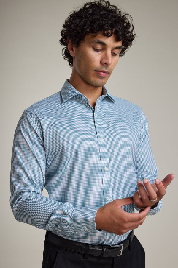 A man with curly dark hair wears the Barkers Zione Business Shirt in light blue and black pants, adjusting his cuff while looking down against a plain light background.